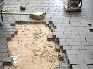 Section of block paving lifted for repair with the sand bed exposed and a plate compactor nearby